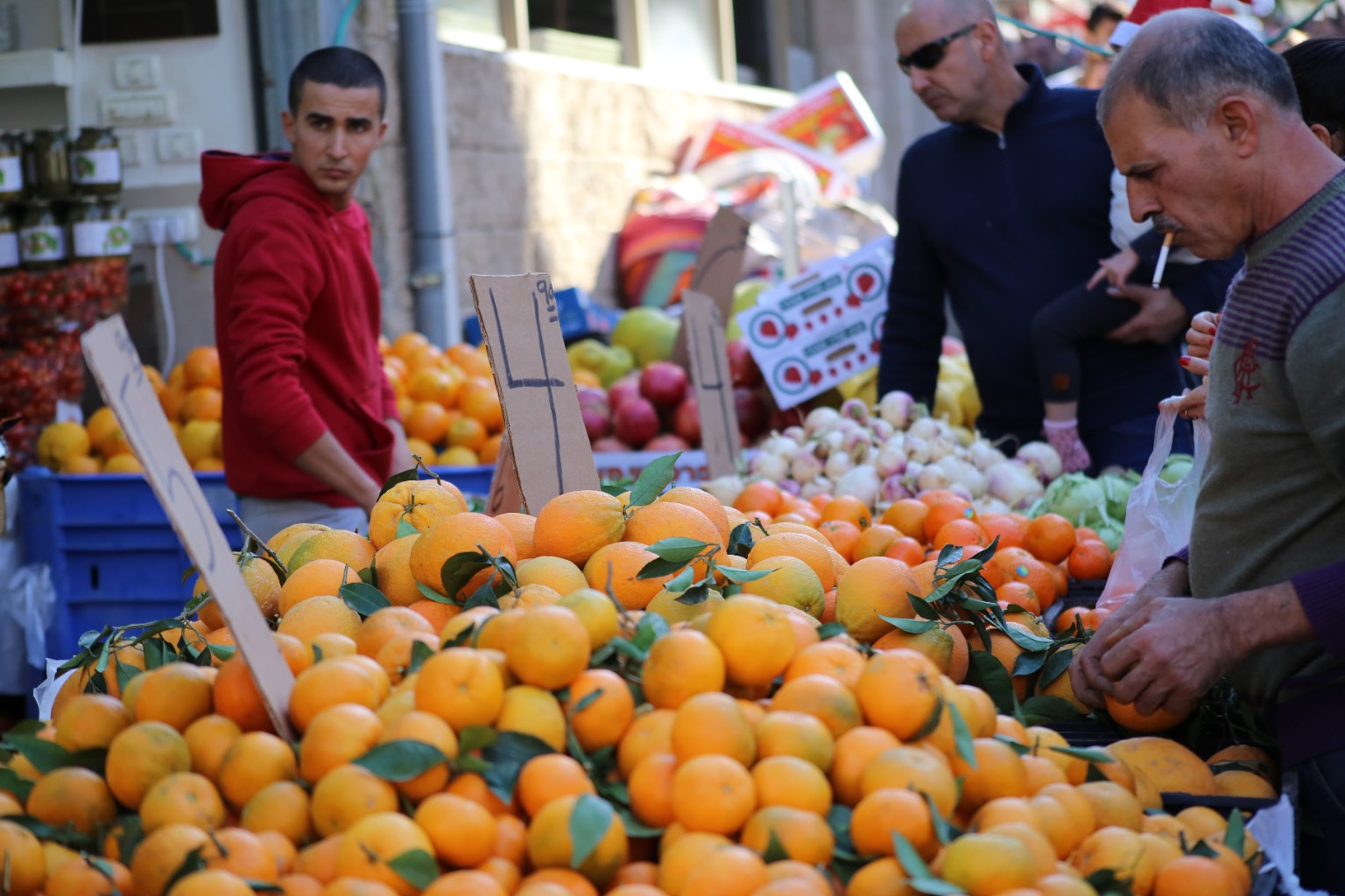 Market stall piled with oranges and green leaves, a man in a red hoodie on the left, blue crates, handwritten cardboard signs, and a gray-haired man selecting fruit on the right.
